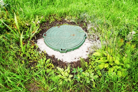 Green plastic septic tank access cover with handles on white concrete riser surrounded by lush grass