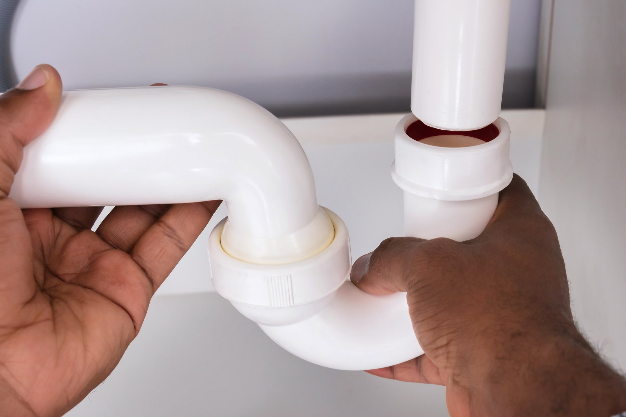 Hands holding a white p-trap under sink during installation or removal of plumbing connections beneath a sink.