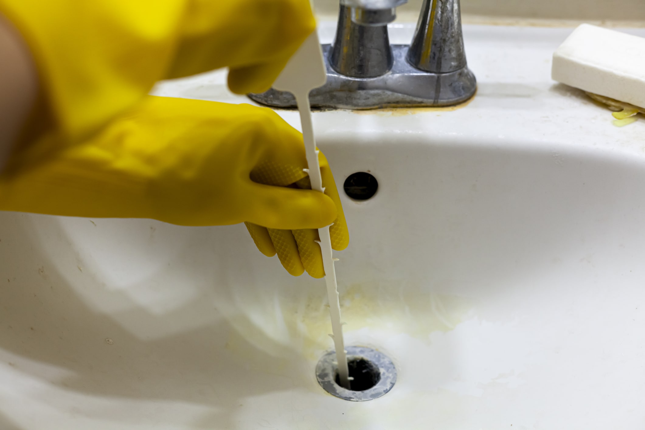A person wearing yellow gloves inserts a plastic drain snake into a sink drain to remove clog inside the drain.